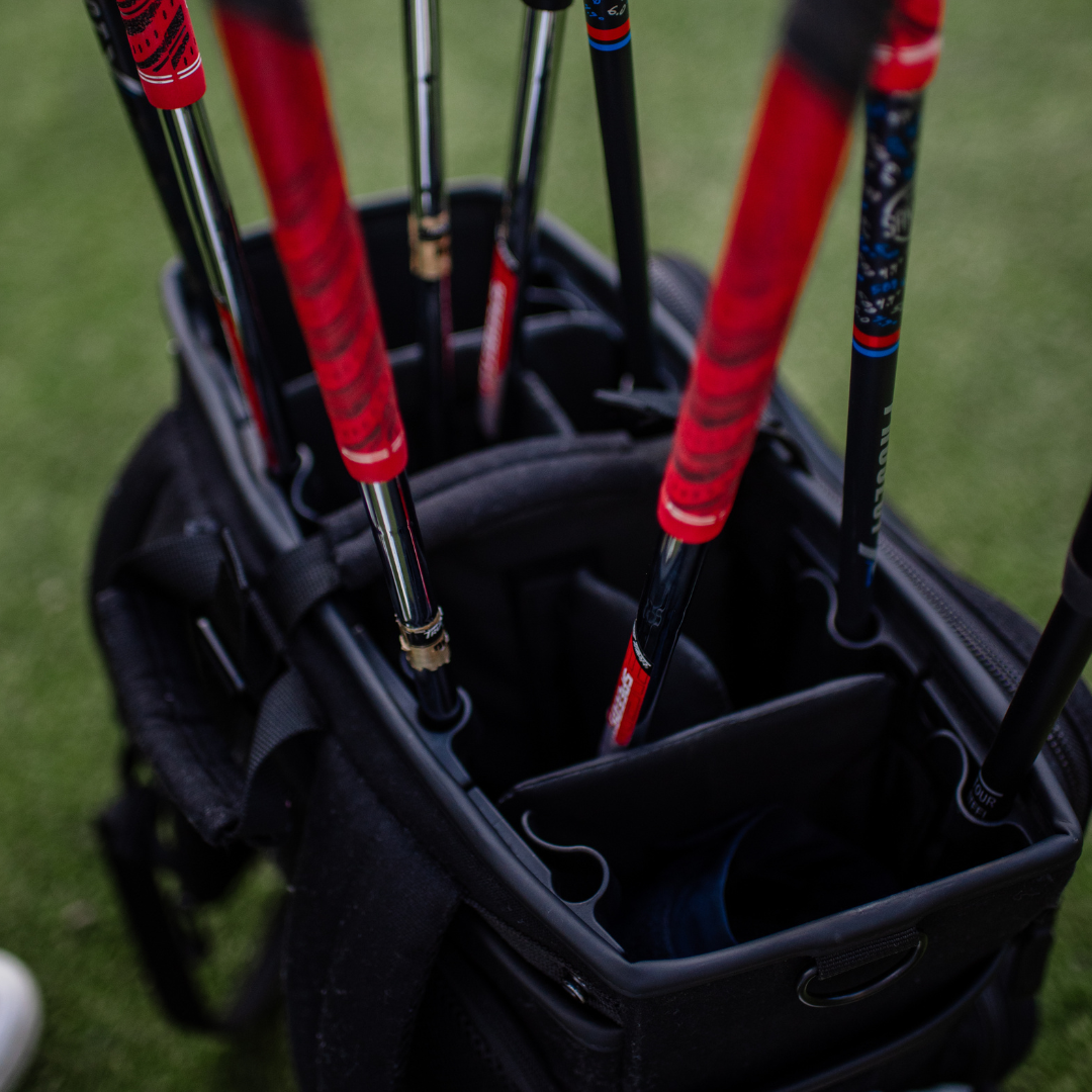 Close-up of golf clubs with red and blue grips in a black bag on a grassy background